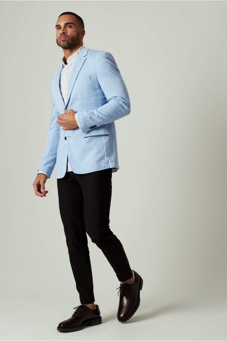 Man wearing a light blue blazer over a white shirt with black pants and shoes on a plain background