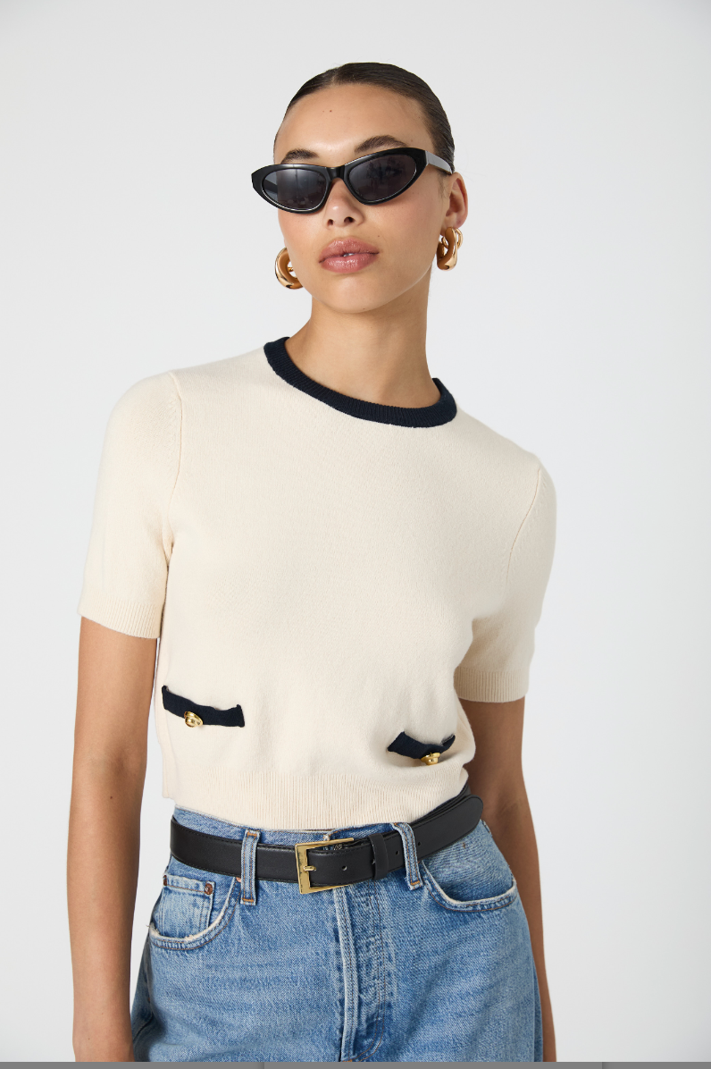Woman wearing a cream sweater with black trim, blue jeans, and sunglasses on a white background