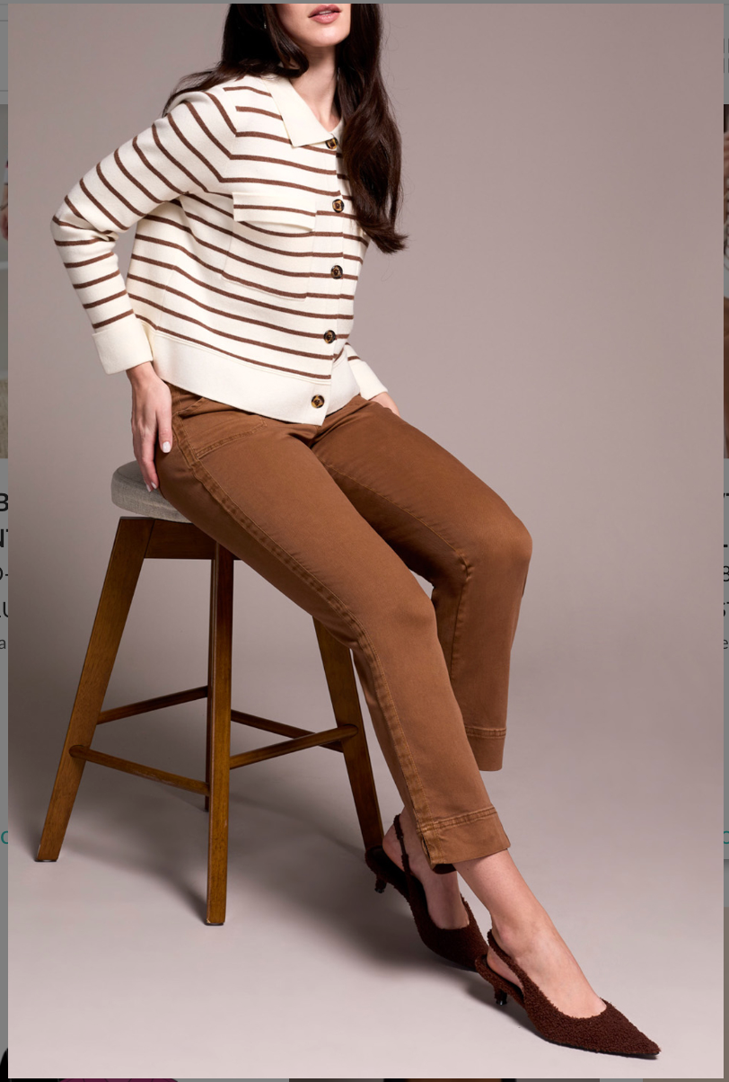 Woman sitting on a stool wearing a striped sweater and brown pants against a plain background