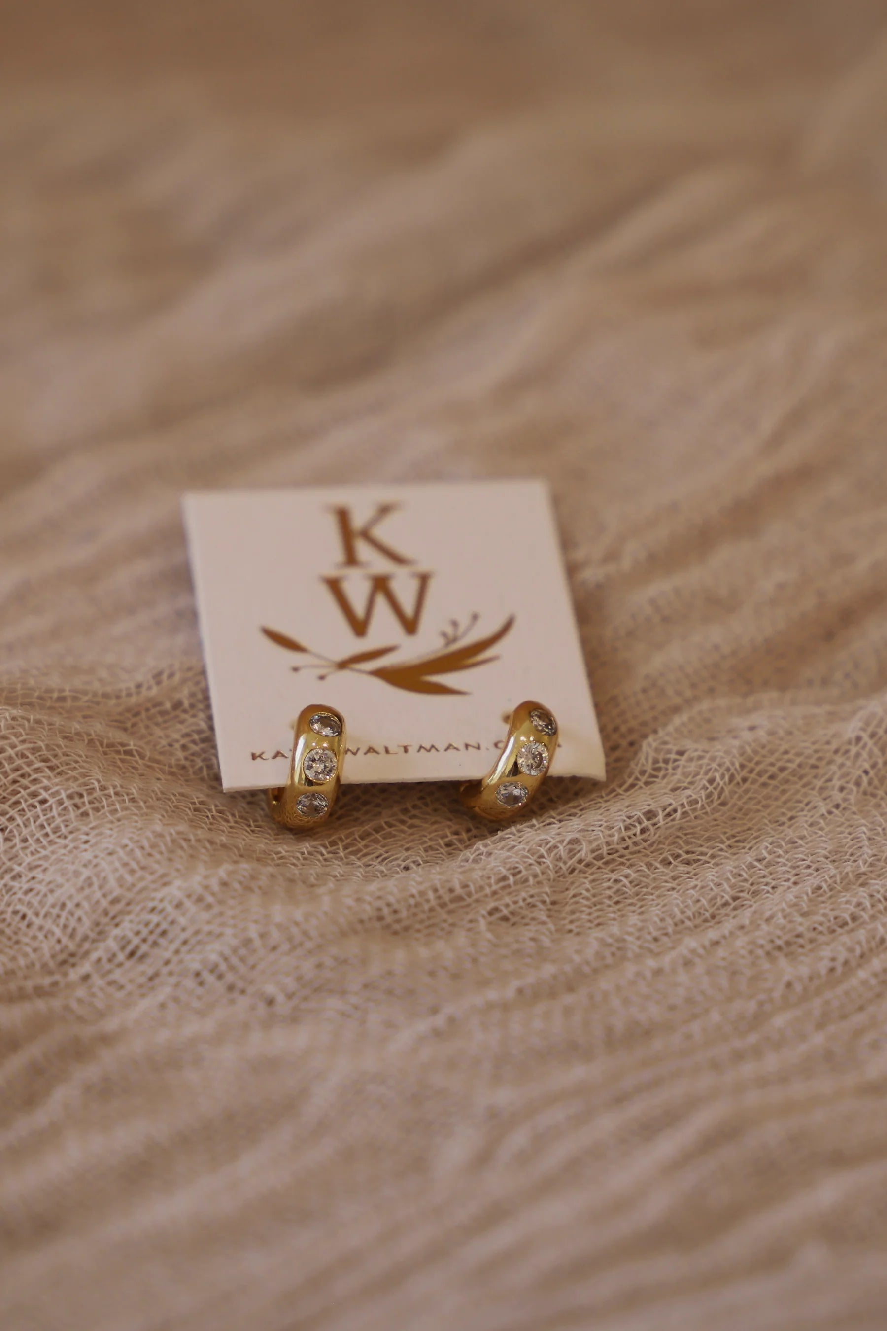 Gold earrings on a wooden display card with 'K W' branding on a textured beige background