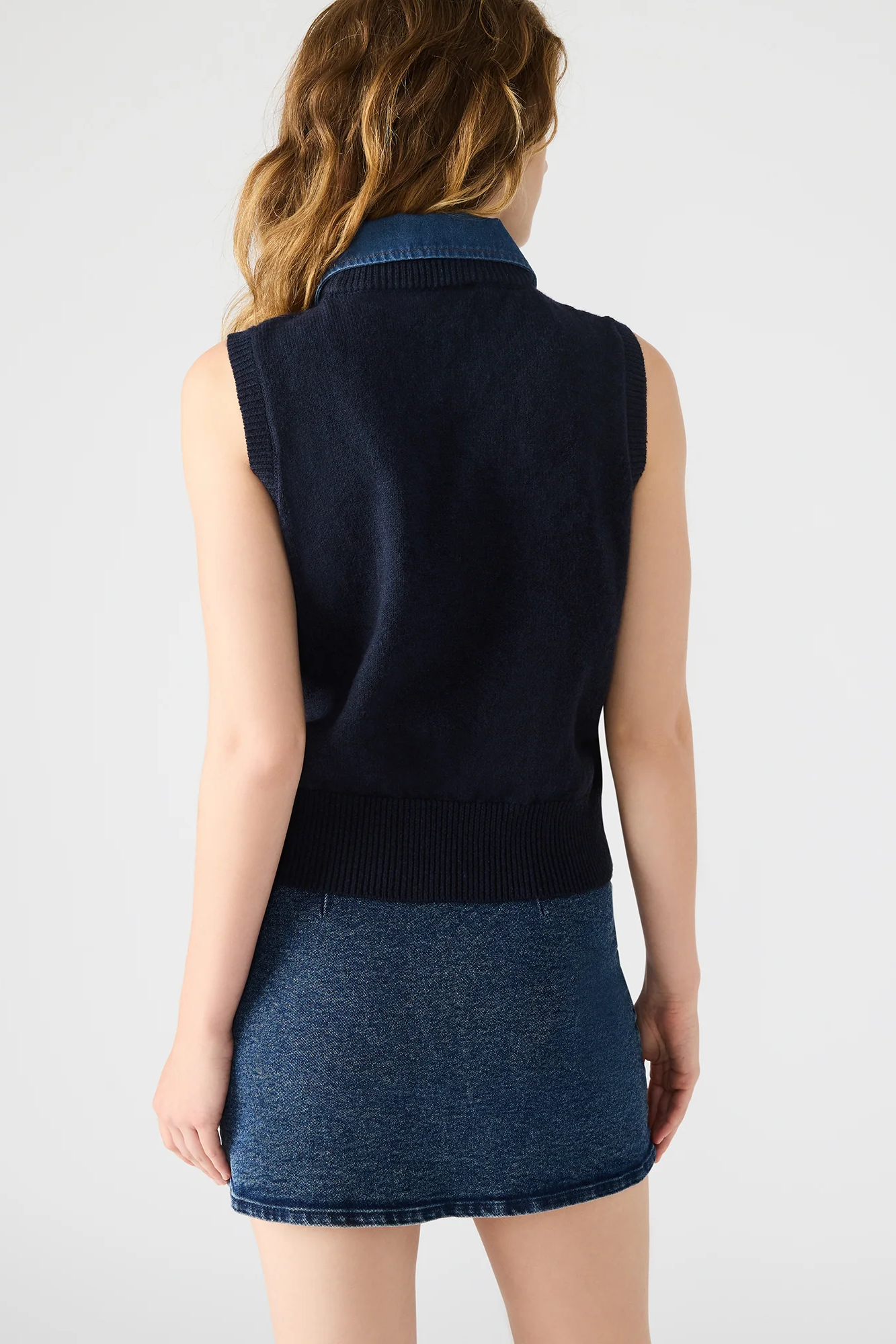 Woman wearing a dark blue sleeveless sweater over a denim skirt on a plain background