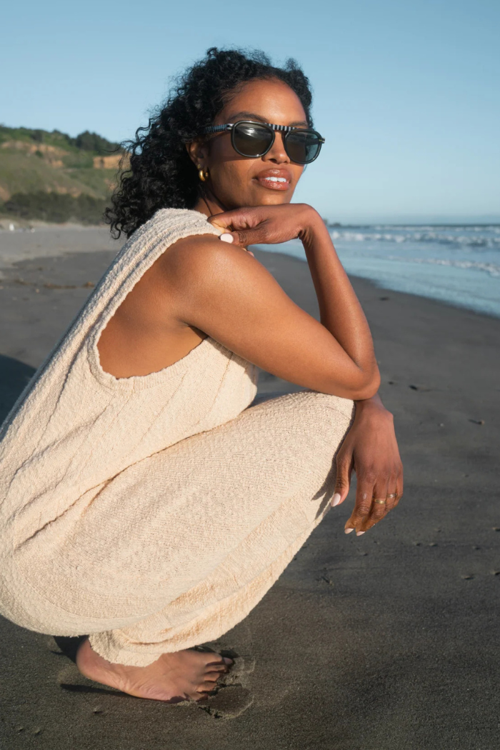 Woman squatting on a beach wearing sunglasses and a beige outfit
