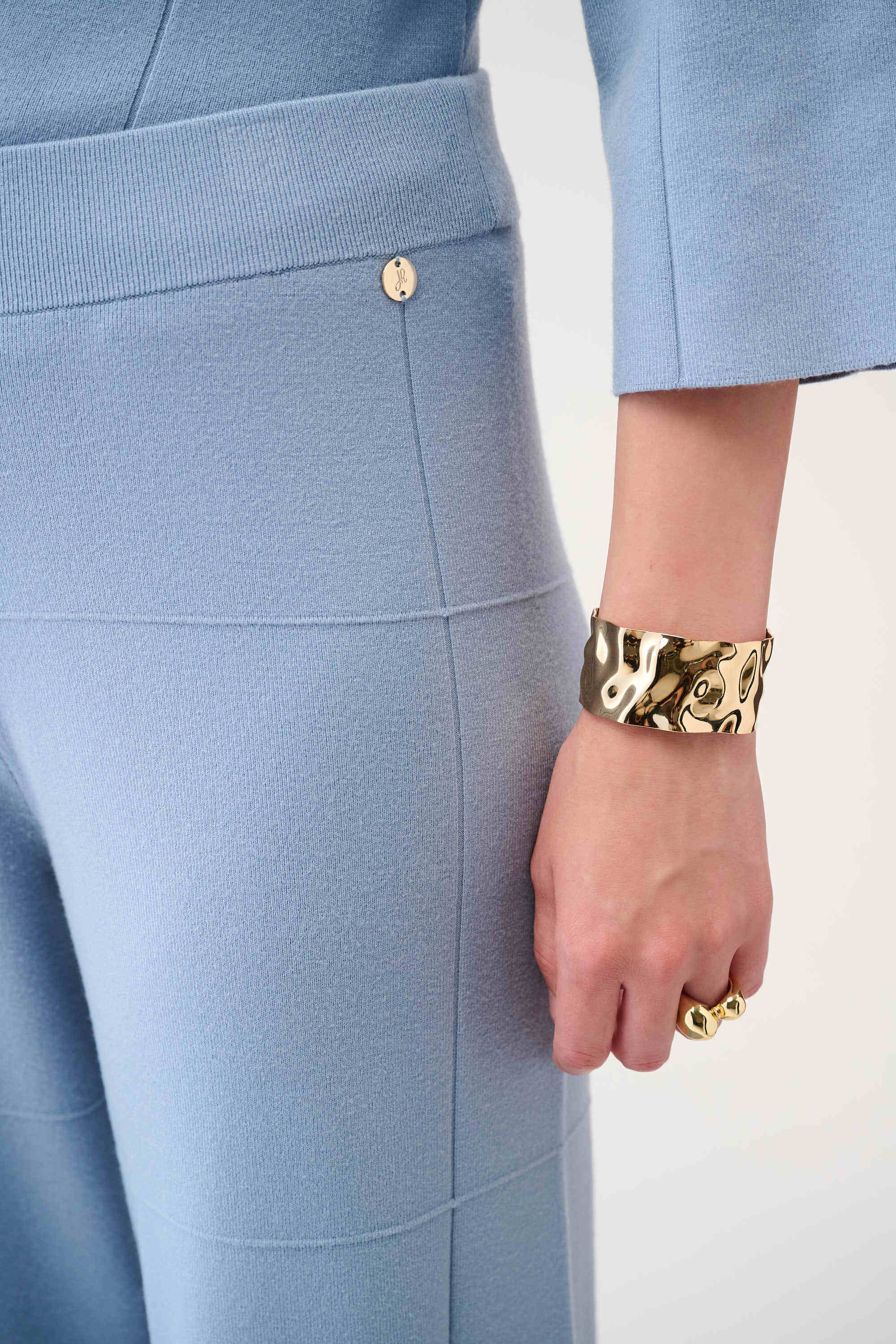 Light blue pants with a gold button and a person wearing a gold bracelet and ring on a white background