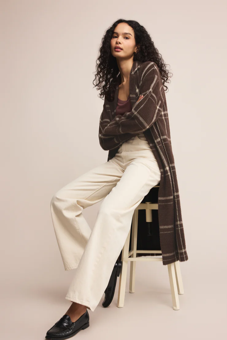 Woman wearing a plaid coat over a white outfit, sitting on a stool against a plain background