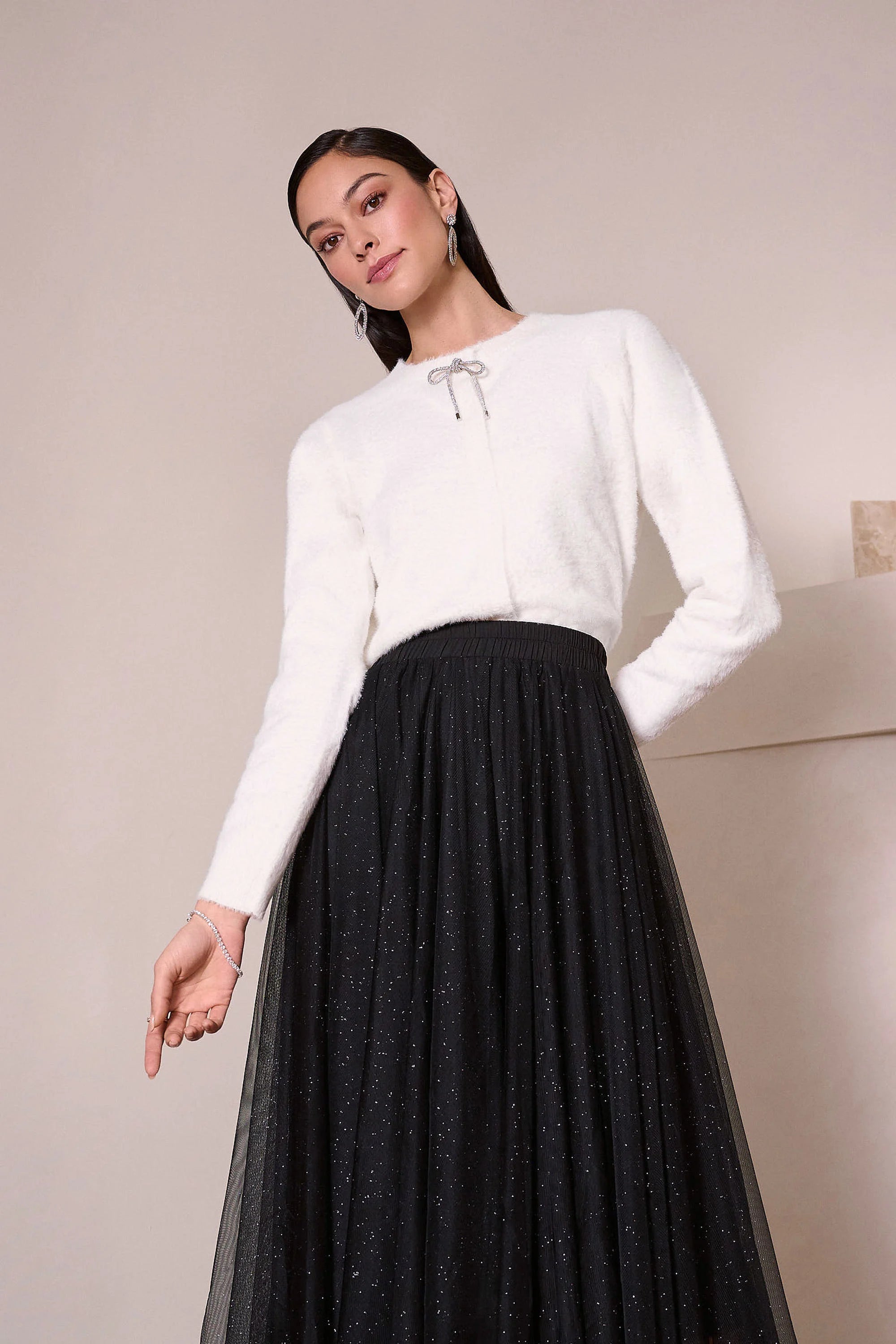 Woman wearing a white long-sleeve top and black pleated skirt against a neutral background