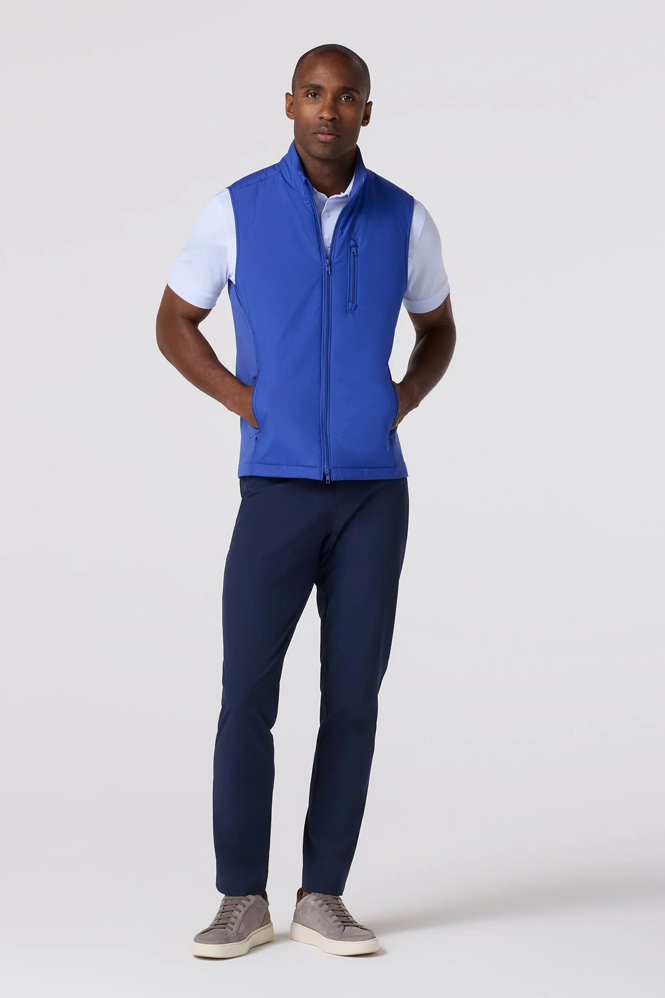 Man wearing a blue vest over a white shirt with navy pants on a white background