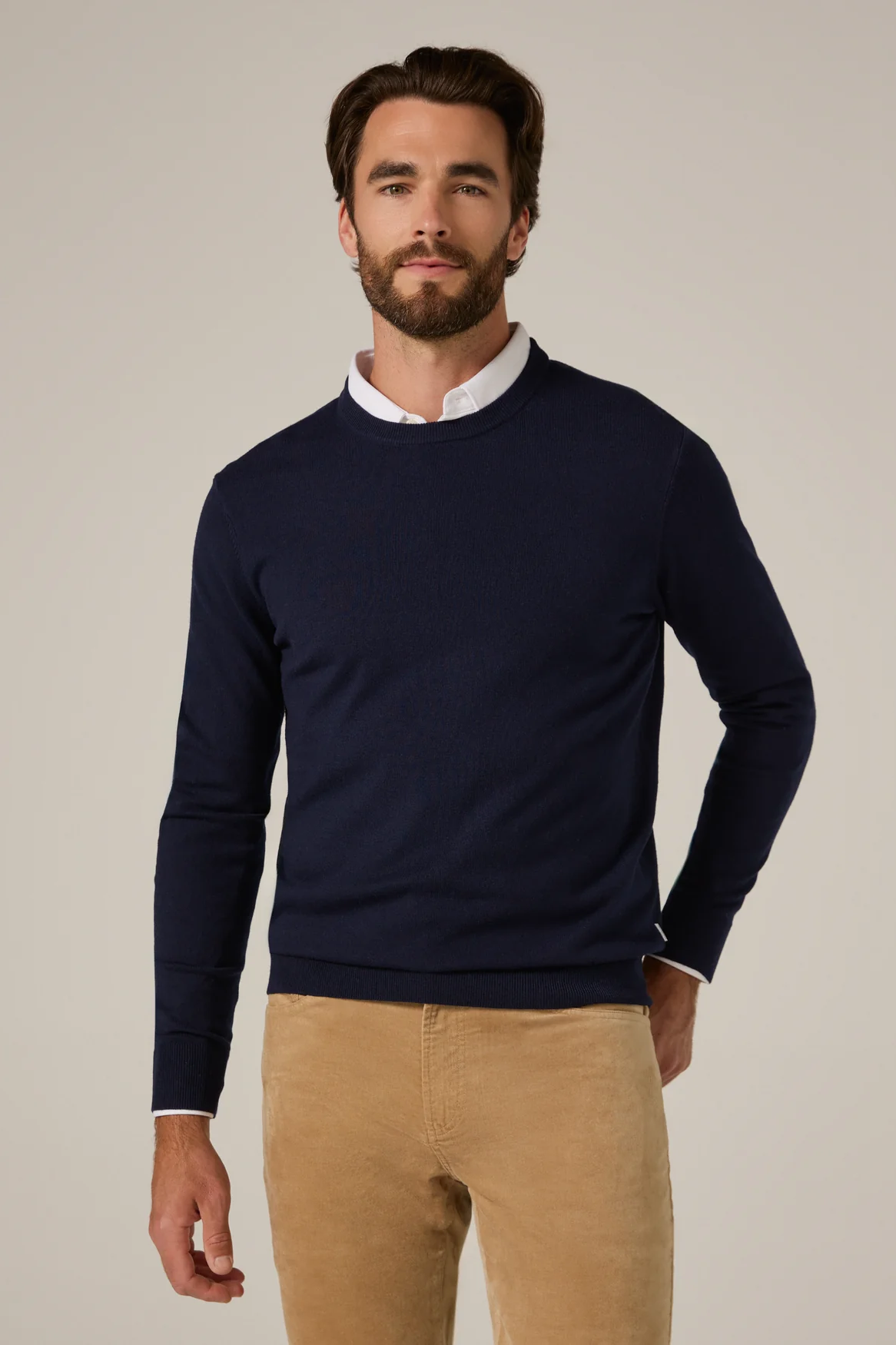 Man wearing a navy sweater over a white shirt with beige pants on a plain background
