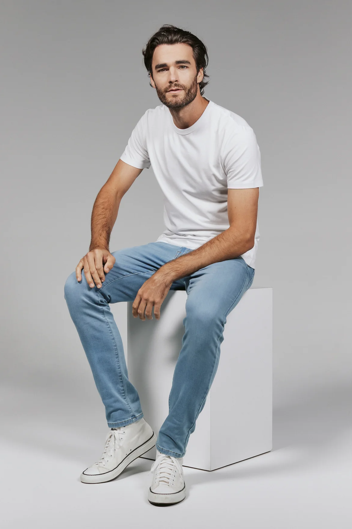 Man sitting on a white cube wearing a white t-shirt and light blue jeans.