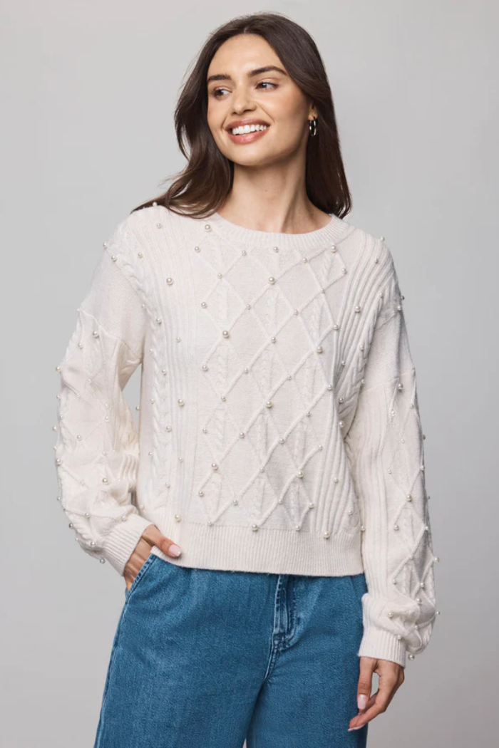Woman wearing a white sweater with pearl embellishments and blue jeans on a gray background