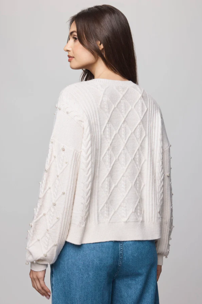Woman wearing a cream-colored cable knit sweater and blue jeans on a gray background