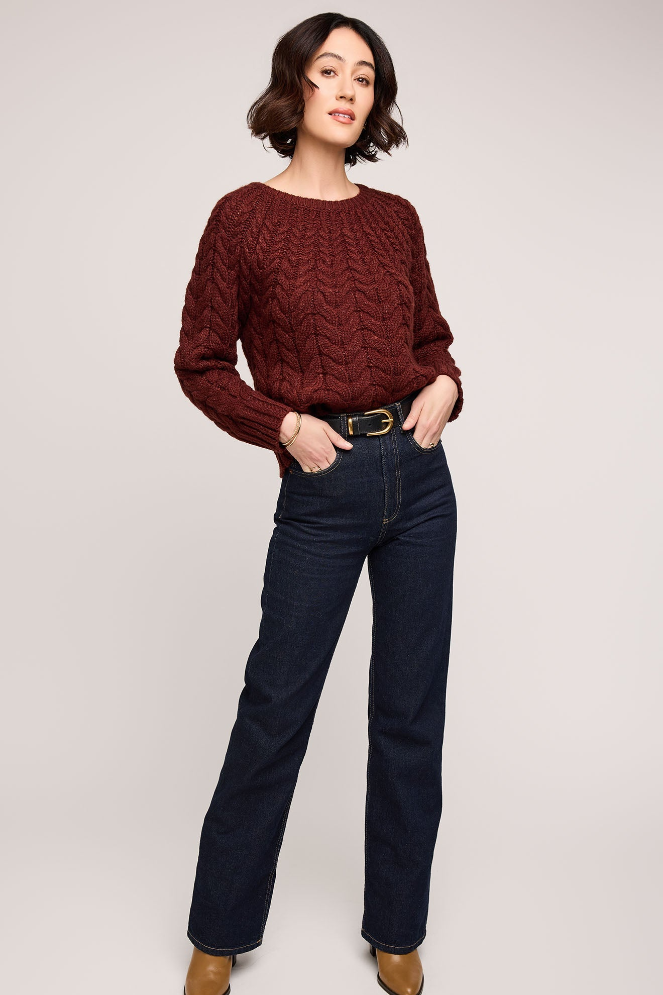 Woman wearing a maroon cable knit sweater and dark blue jeans on a plain background