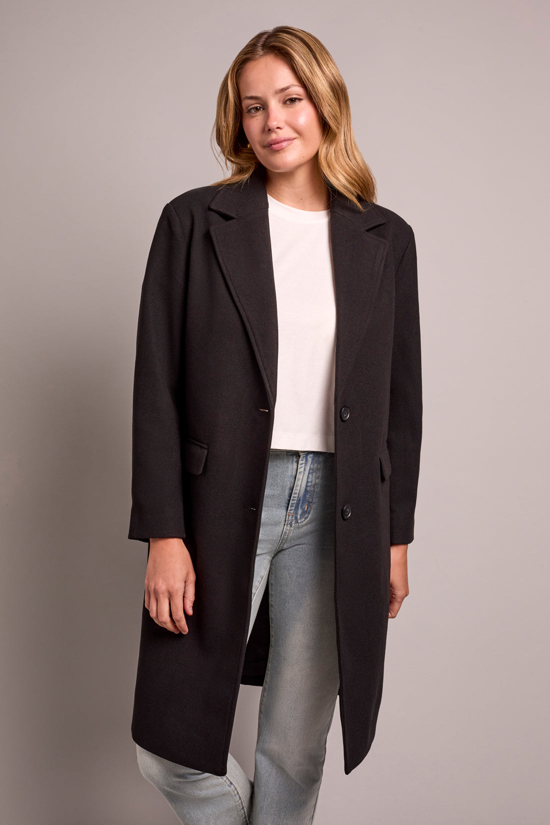 Woman wearing a black coat over a white shirt and jeans on a gray background