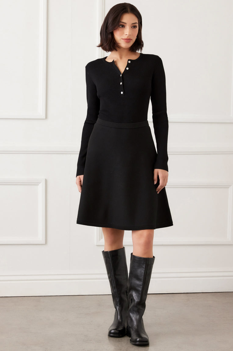 Woman wearing a black dress with long sleeves and a skirt, standing against a white wall.