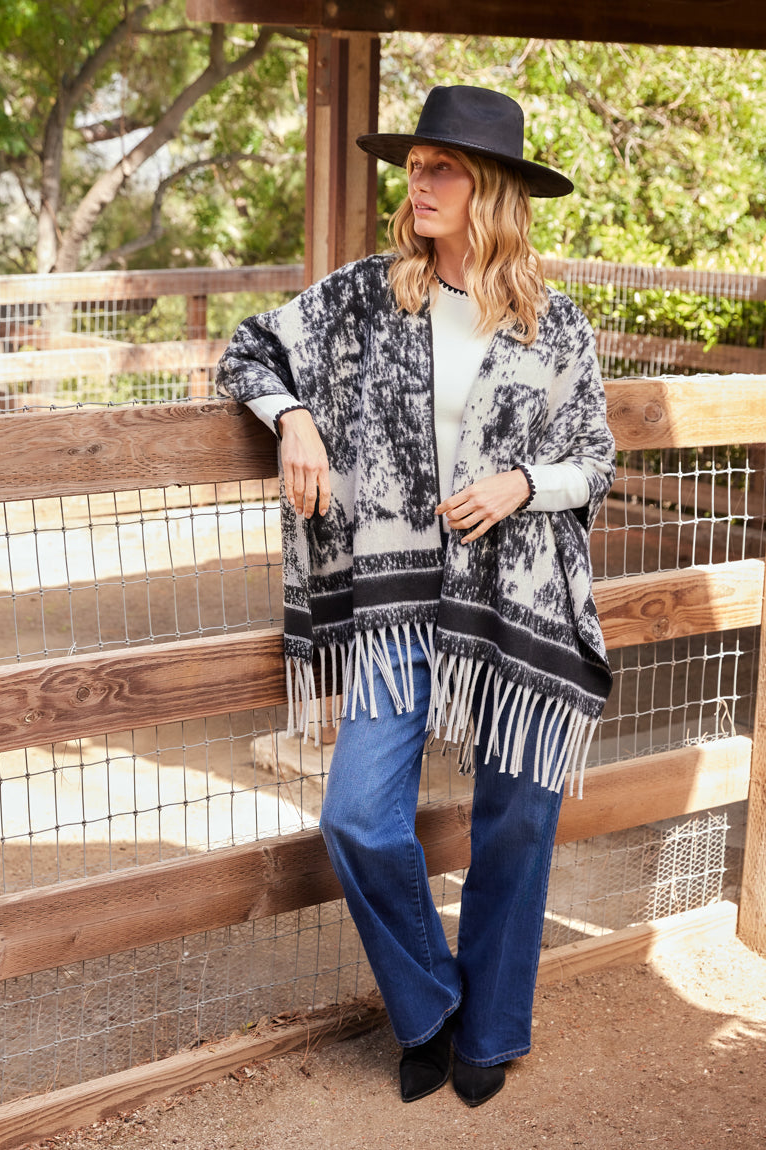 Woman wearing a black and white patterned poncho with fringe, blue jeans, and a black hat, standing by a wooden fence.