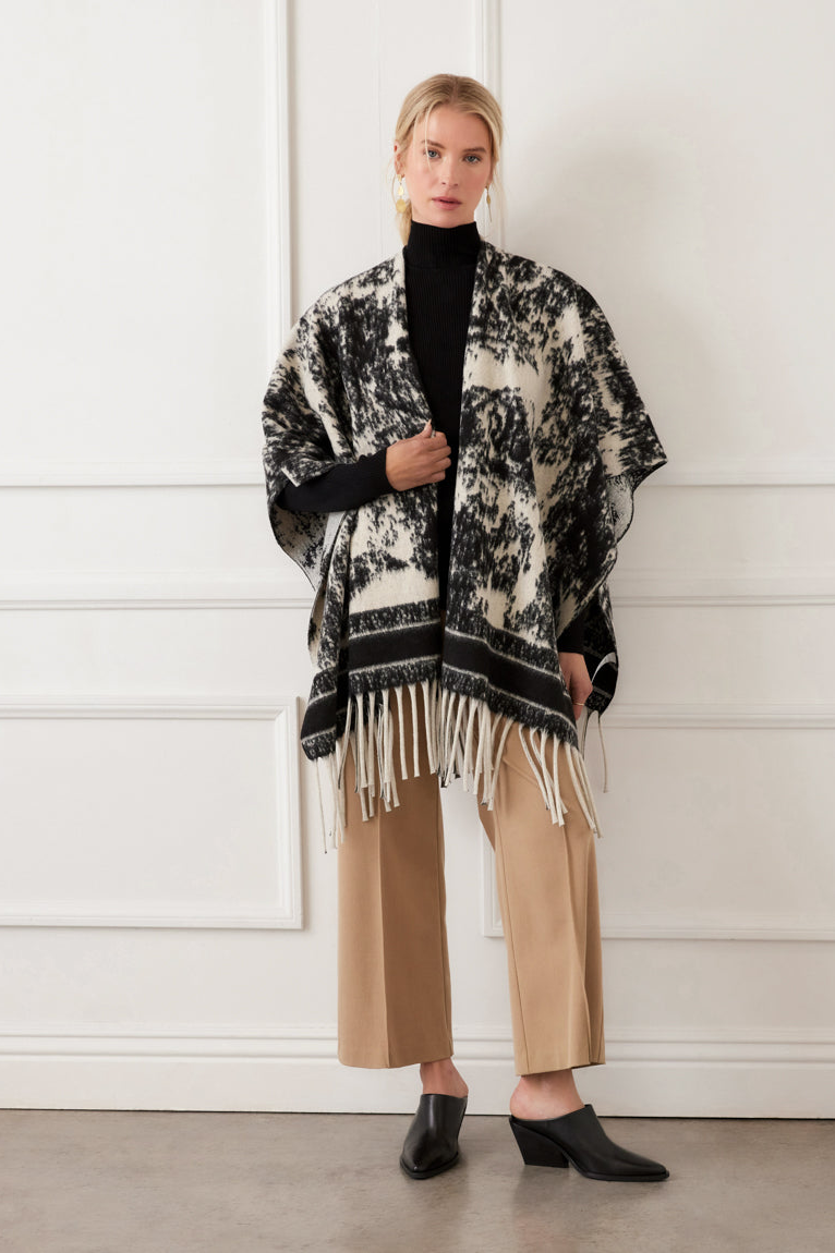 Woman wearing a black and white patterned poncho with fringe, black turtleneck, beige pants, and black shoes against a white background.