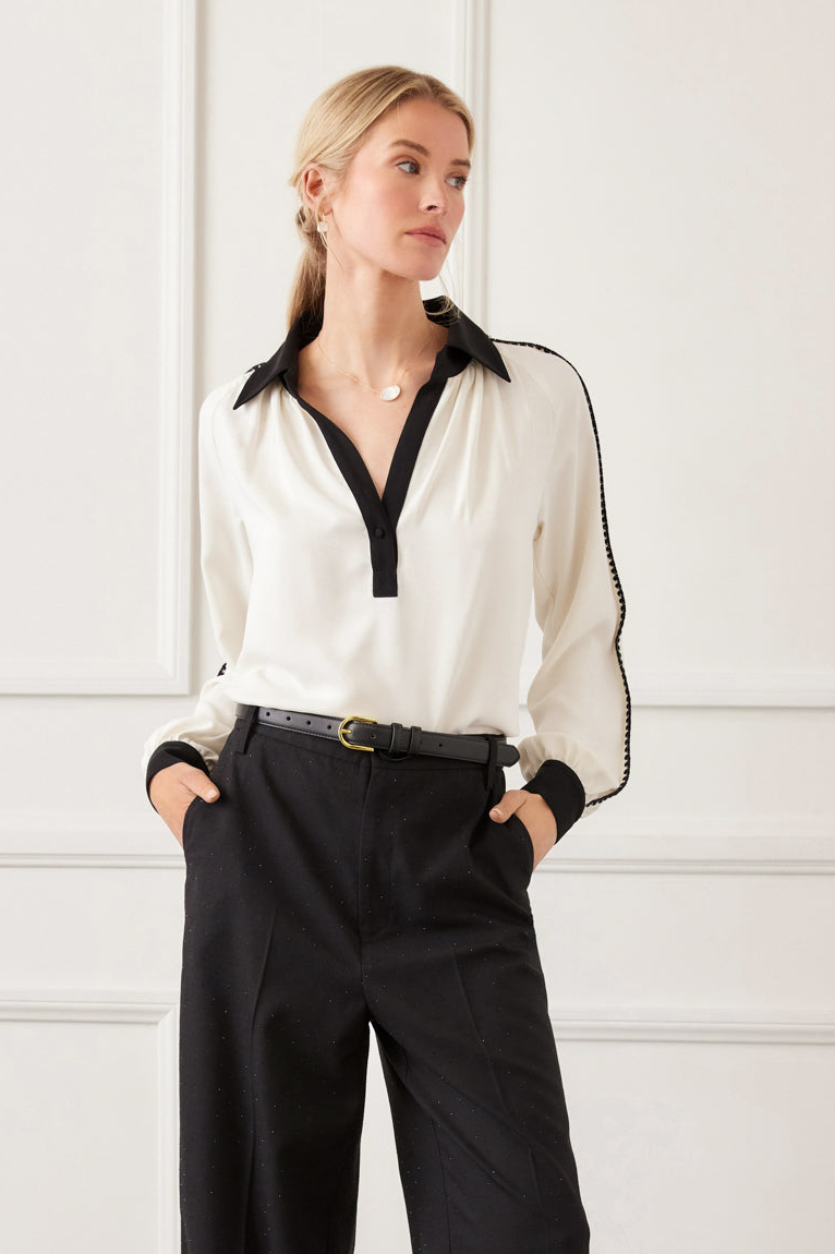 Woman wearing a white blouse with black accents and black pants against a white wall.