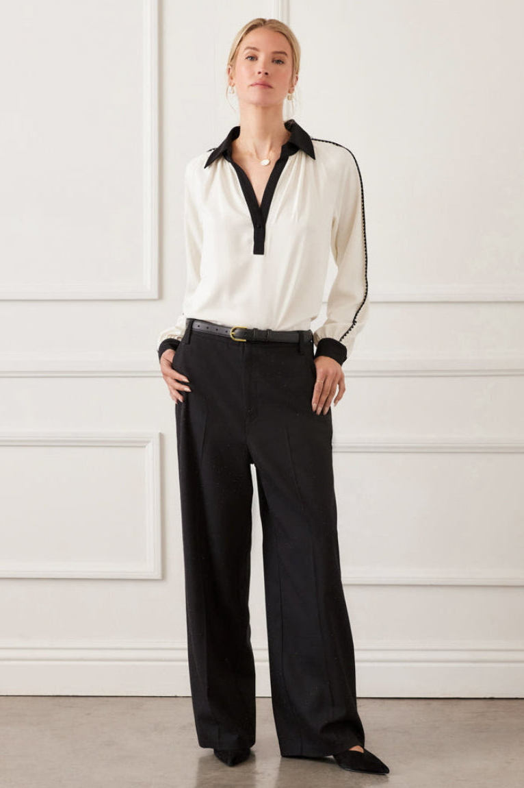 Woman wearing a white blouse with black trim and black pants against a white wall.