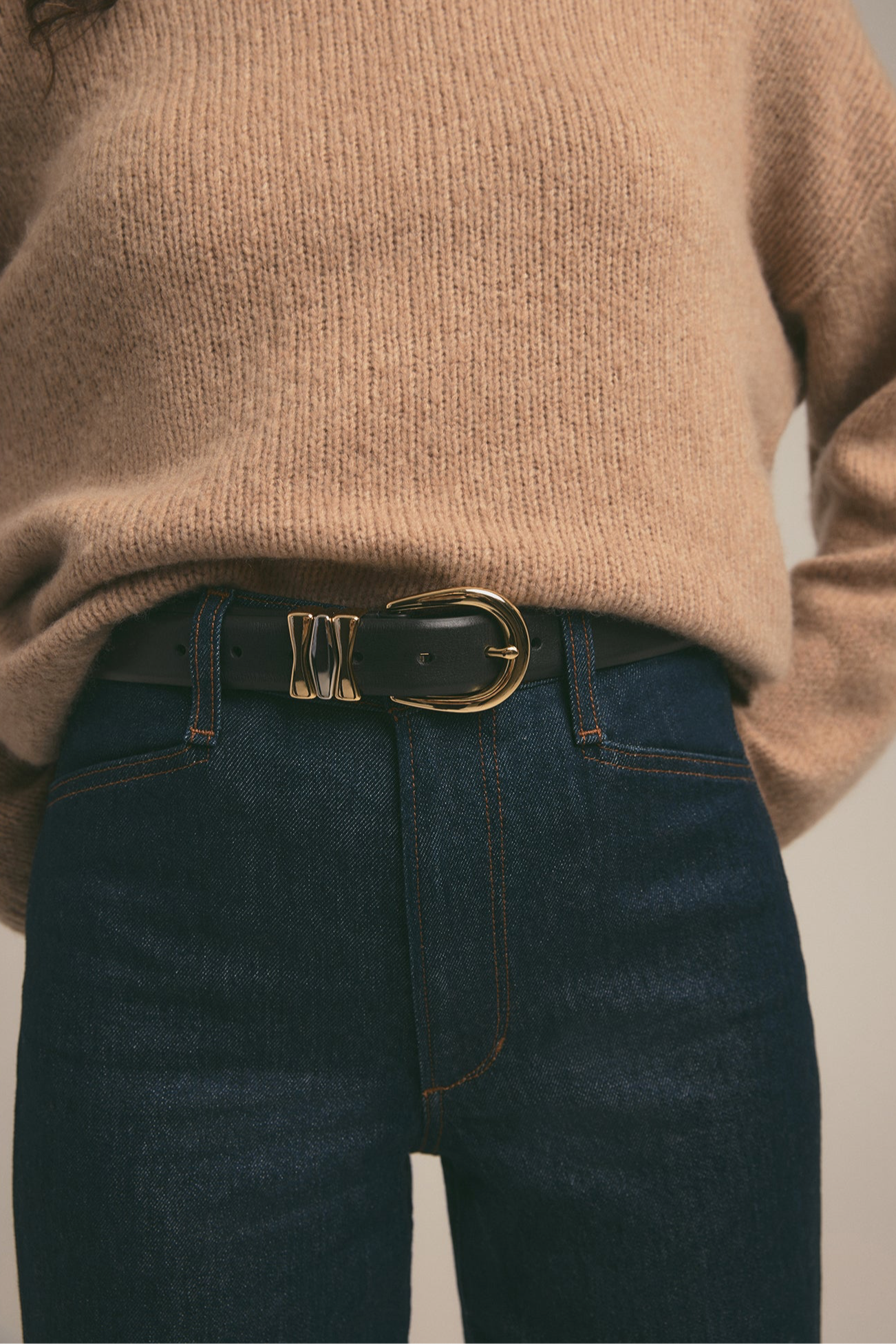 Person wearing a beige sweater and dark blue jeans with a gold belt buckle.