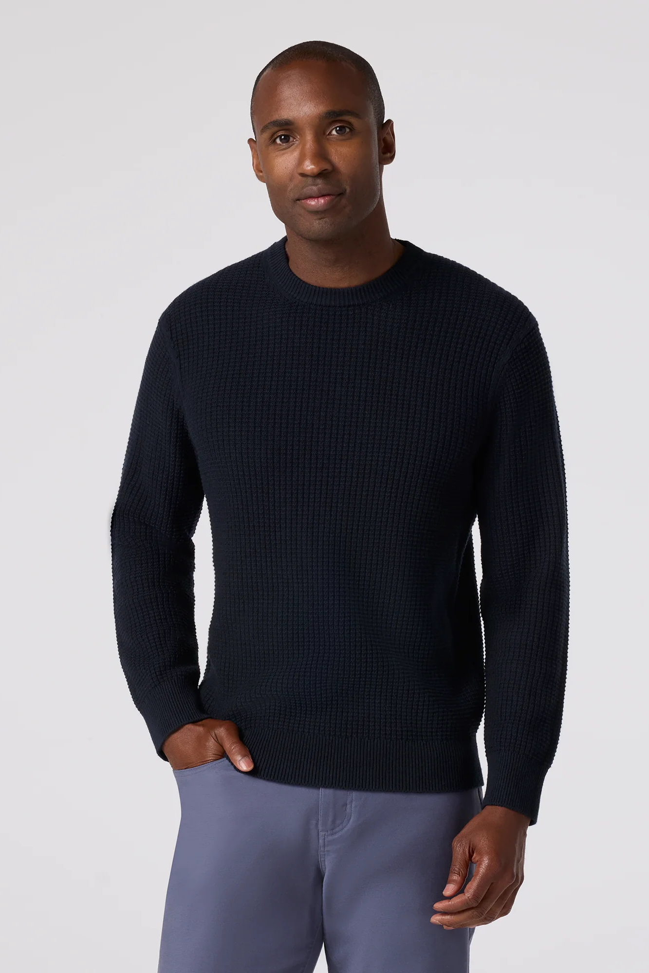Man wearing a dark blue sweater and light gray pants against a white background