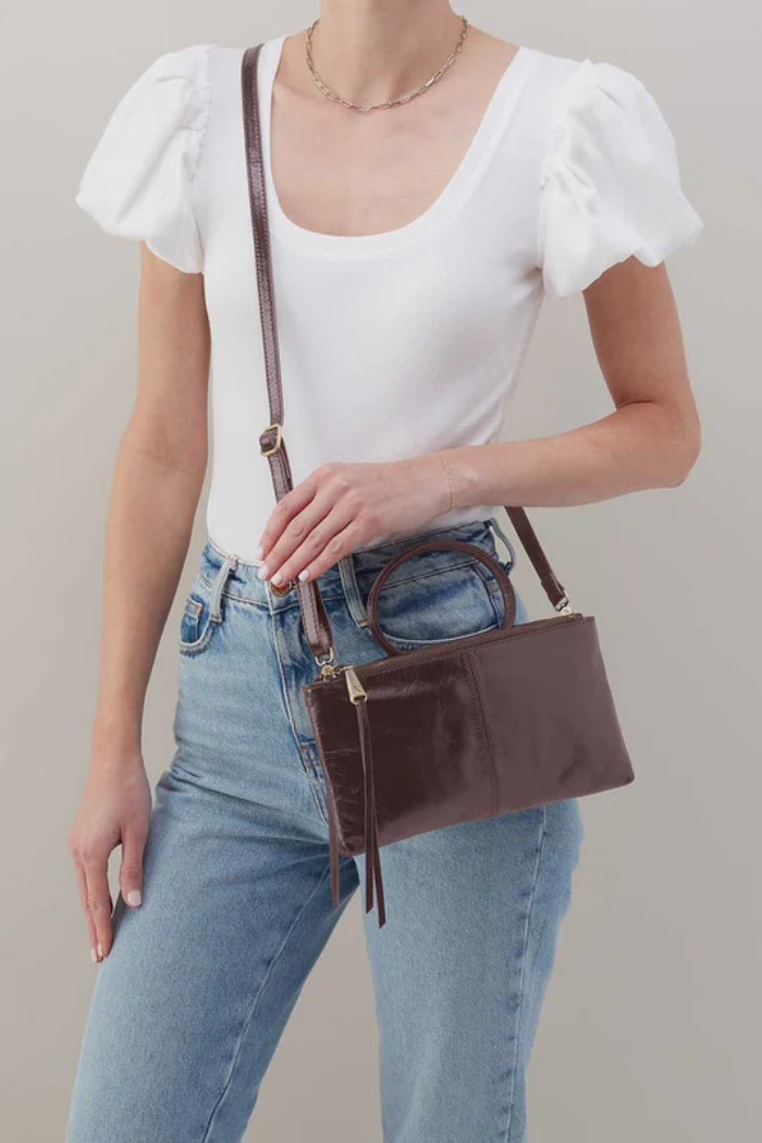 Person wearing a white top and blue jeans holding a brown handbag against a plain background
