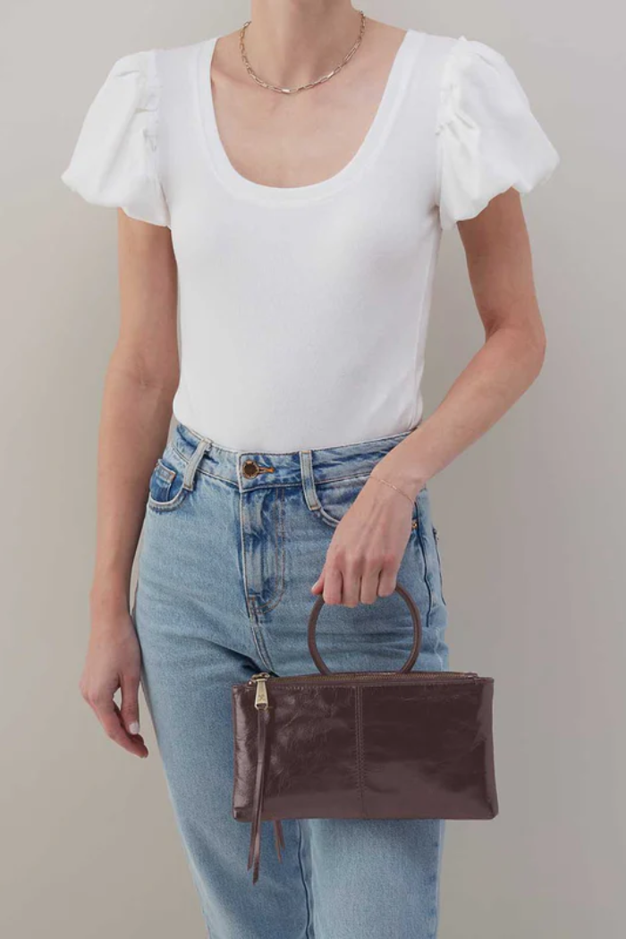 Person wearing a white top and blue jeans holding a brown leather handbag against a neutral background