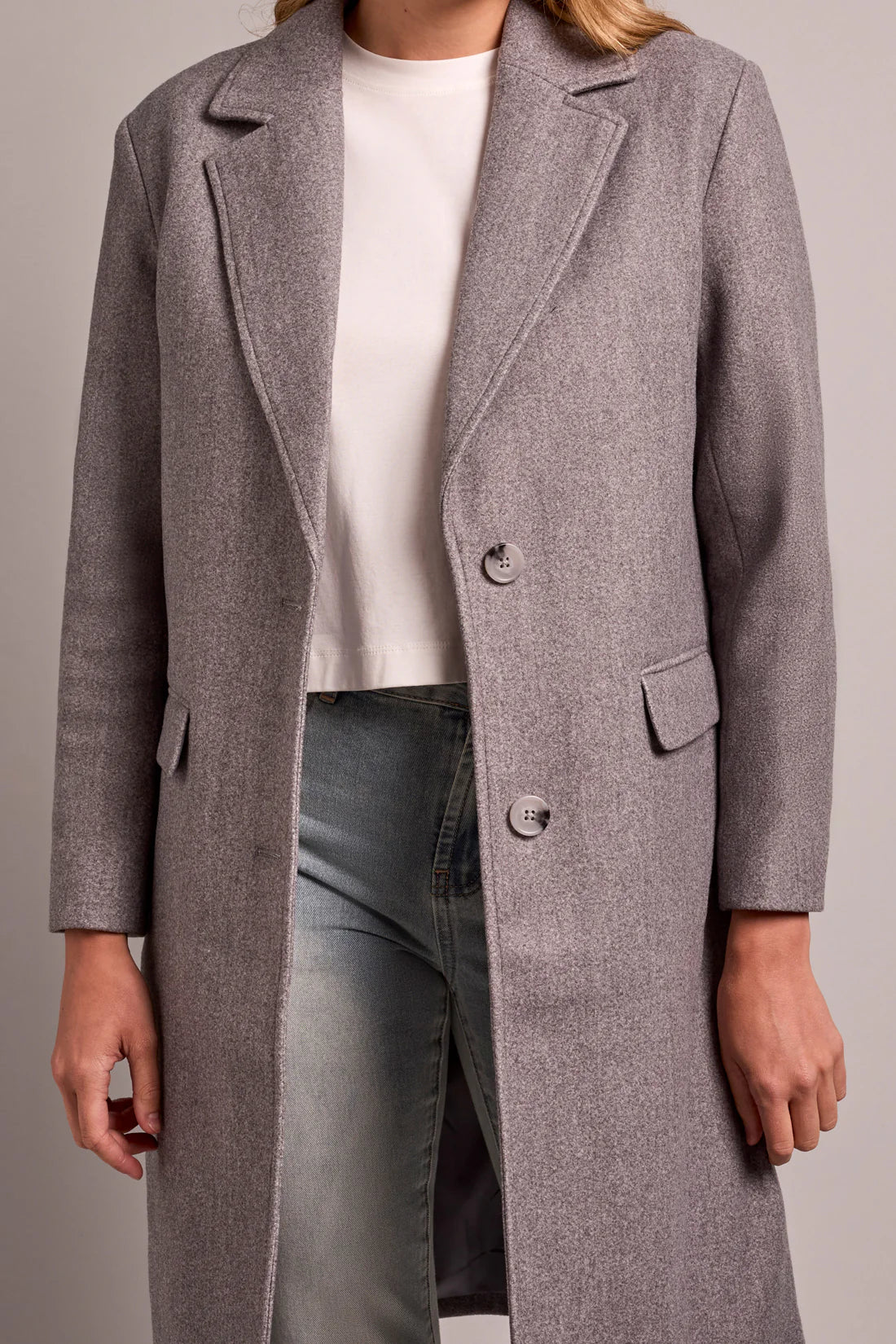 Person wearing a gray coat over a white shirt and dark jeans on a plain background