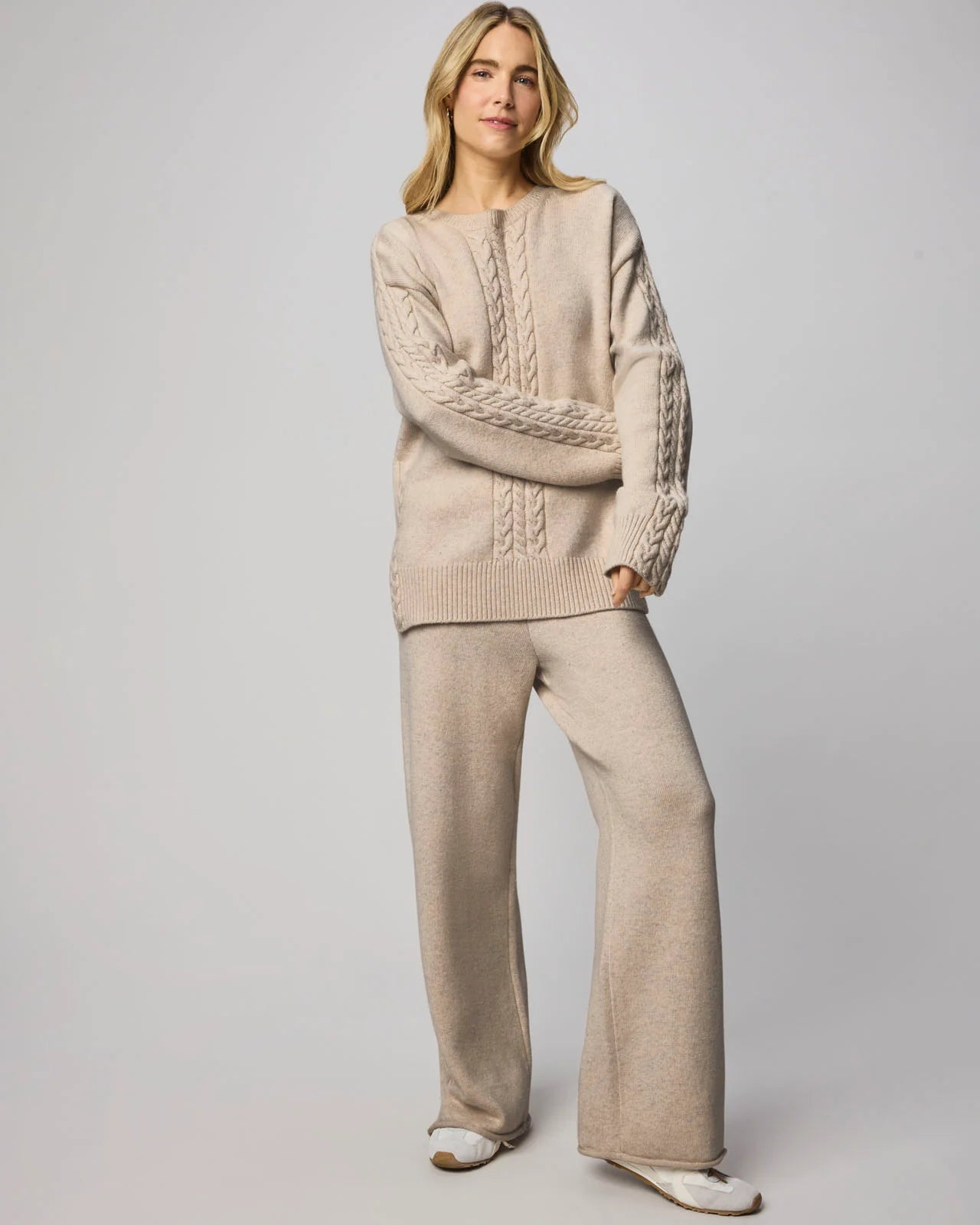 Woman wearing a beige cable knit sweater and matching pants on a plain background
