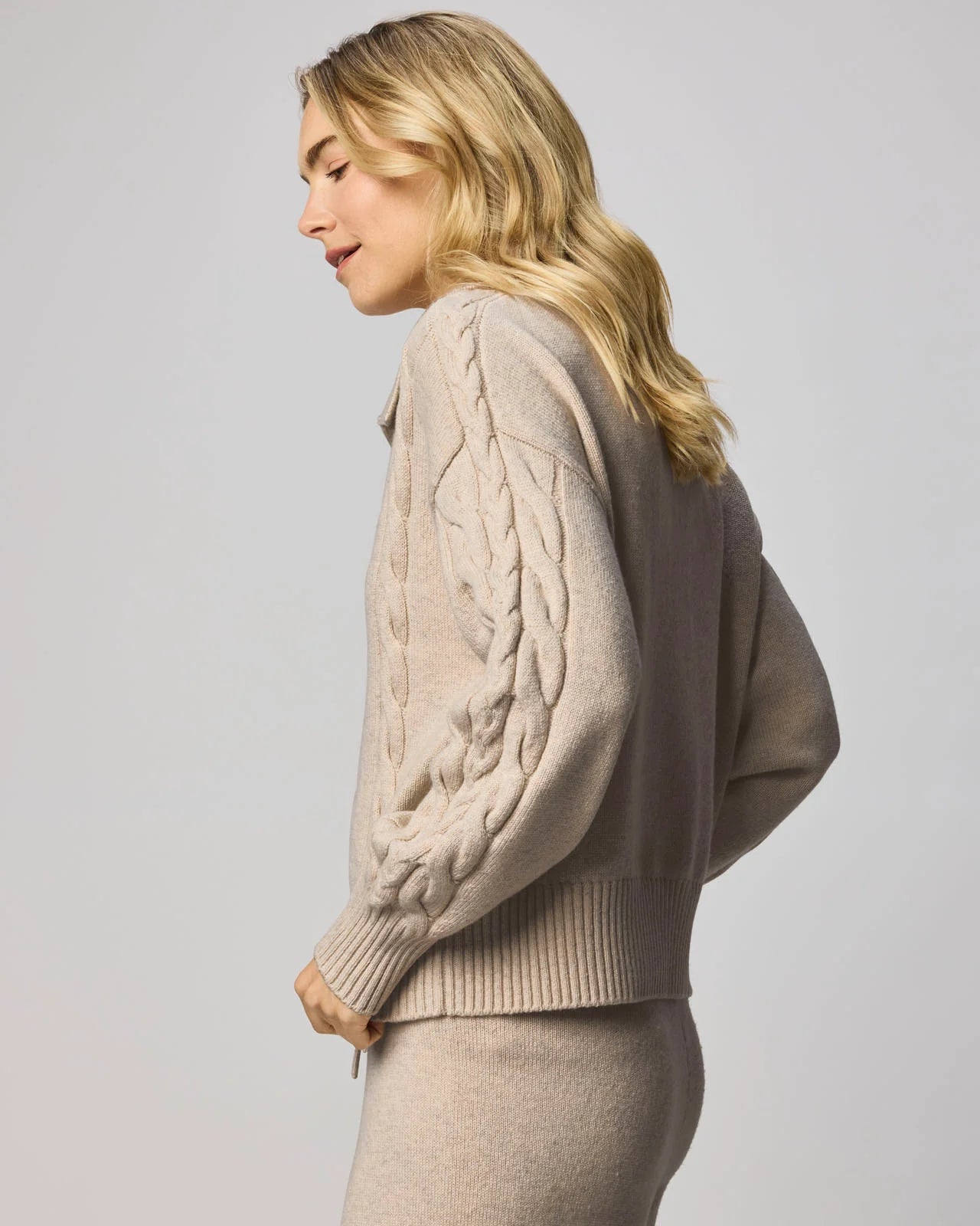 Woman wearing a beige cable knit sweater against a plain background