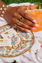 Hand with jewelry holding a glass of orange juice next to a decorative plate with cards on a white surface.