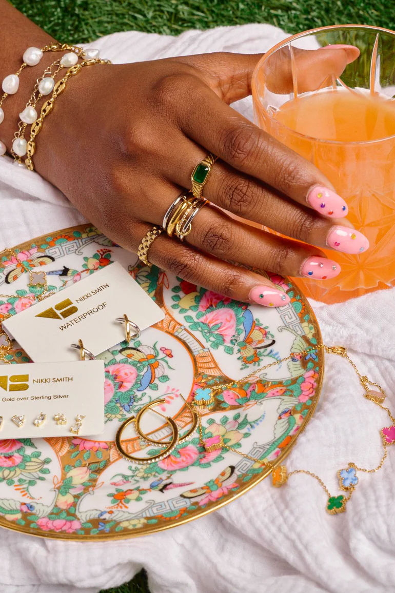 Hand with jewelry holding a glass of orange juice next to a decorative plate with cards on a white surface.