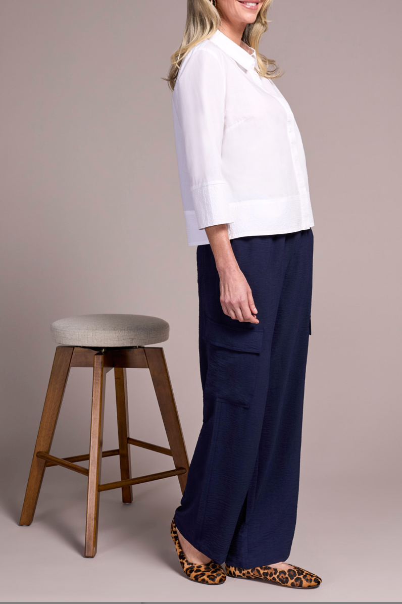 Woman wearing a white blouse and navy pants standing next to a wooden stool on a plain background