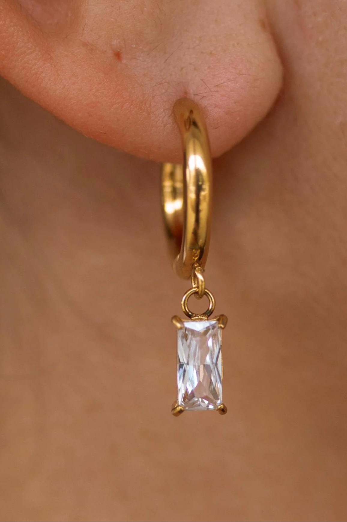 Gold hoop earring with a clear gemstone on a person's ear.