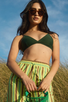 Woman wearing a green bikini top and striped skirt in a grassy field with a blue sky.