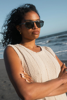 Woman wearing sunglasses and a beige dress standing on a beach with ocean in the background