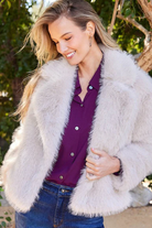 Woman wearing a white fur coat over a purple shirt with a blurred natural background