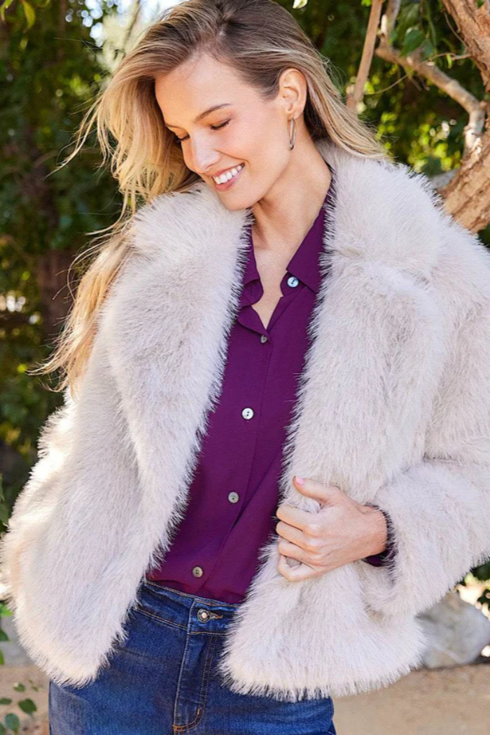 Woman wearing a white fur coat over a purple shirt with a blurred natural background