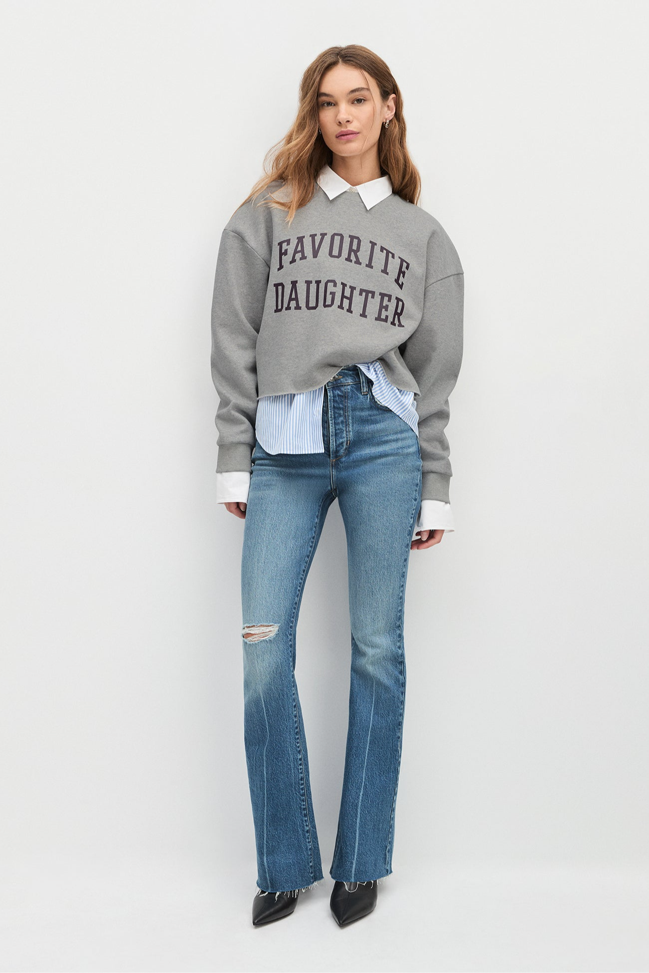 Woman wearing a gray sweatshirt with 'Favorite Daughter' text, white shirt, and blue jeans on a white background