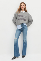Woman wearing a gray sweatshirt with 'Favorite Daughter' text, white shirt, and blue jeans on a white background