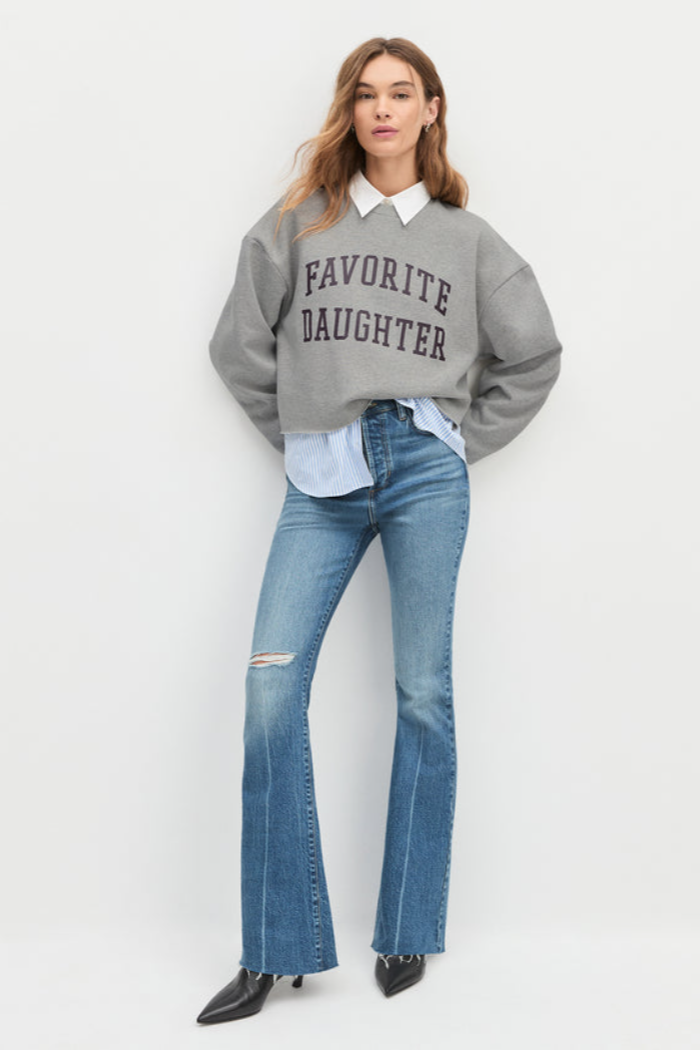 Woman wearing a gray sweatshirt with 'Favorite Daughter' text, white shirt, and blue jeans on a white background