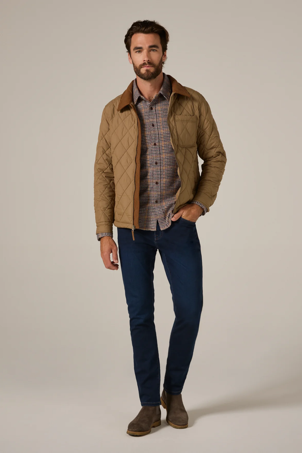 Man wearing a quilted jacket, plaid shirt, and jeans on a plain background