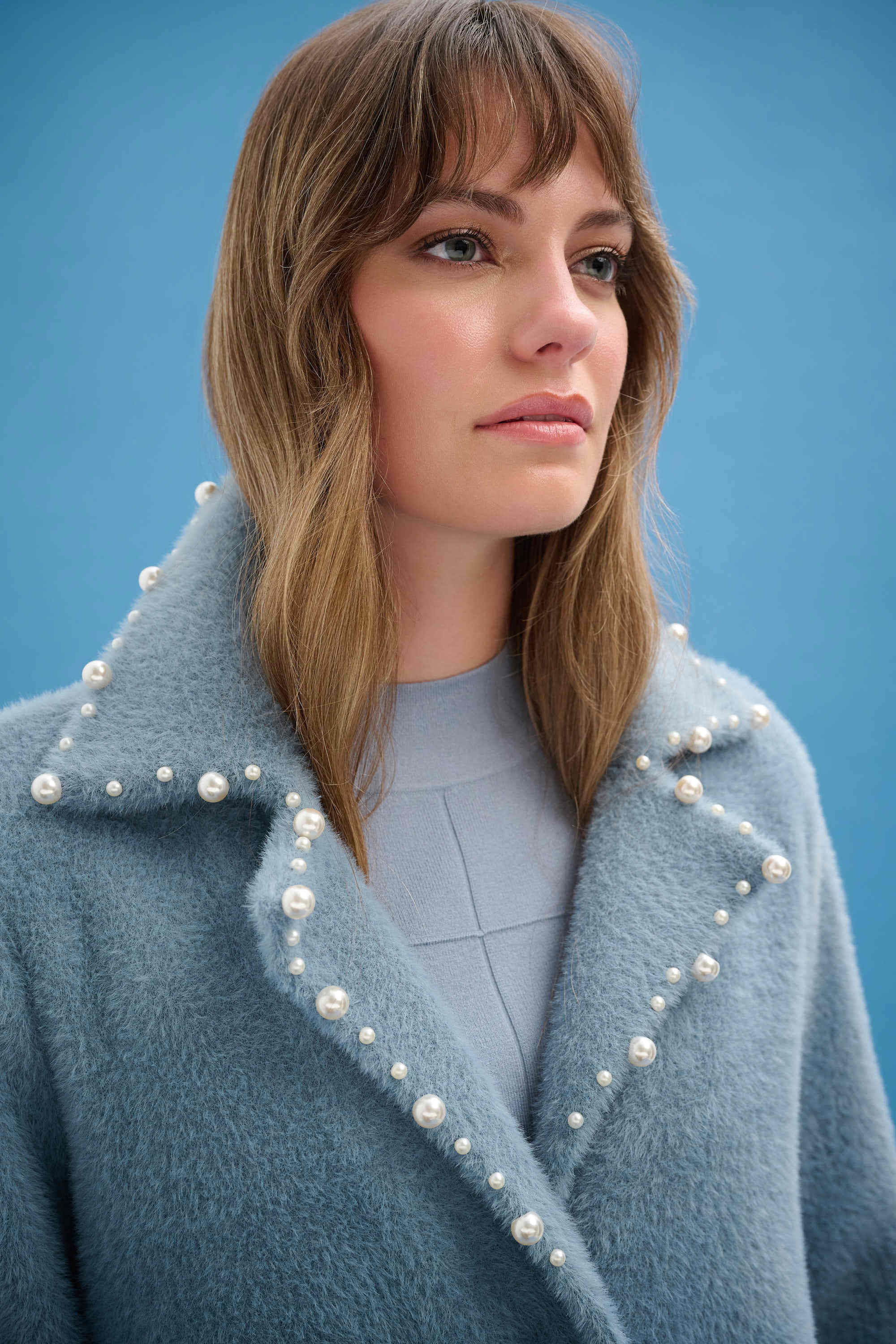 Woman wearing a blue coat with pearl embellishments against a blue background