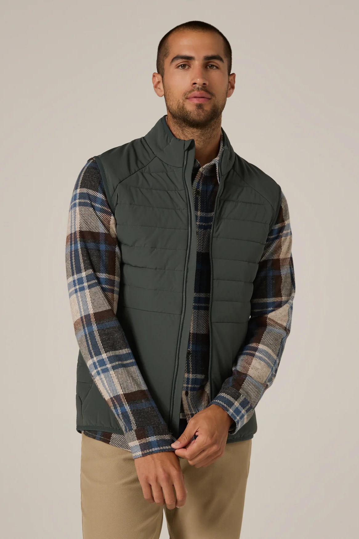 Man wearing a green quilted vest over a plaid shirt with beige pants on a plain background