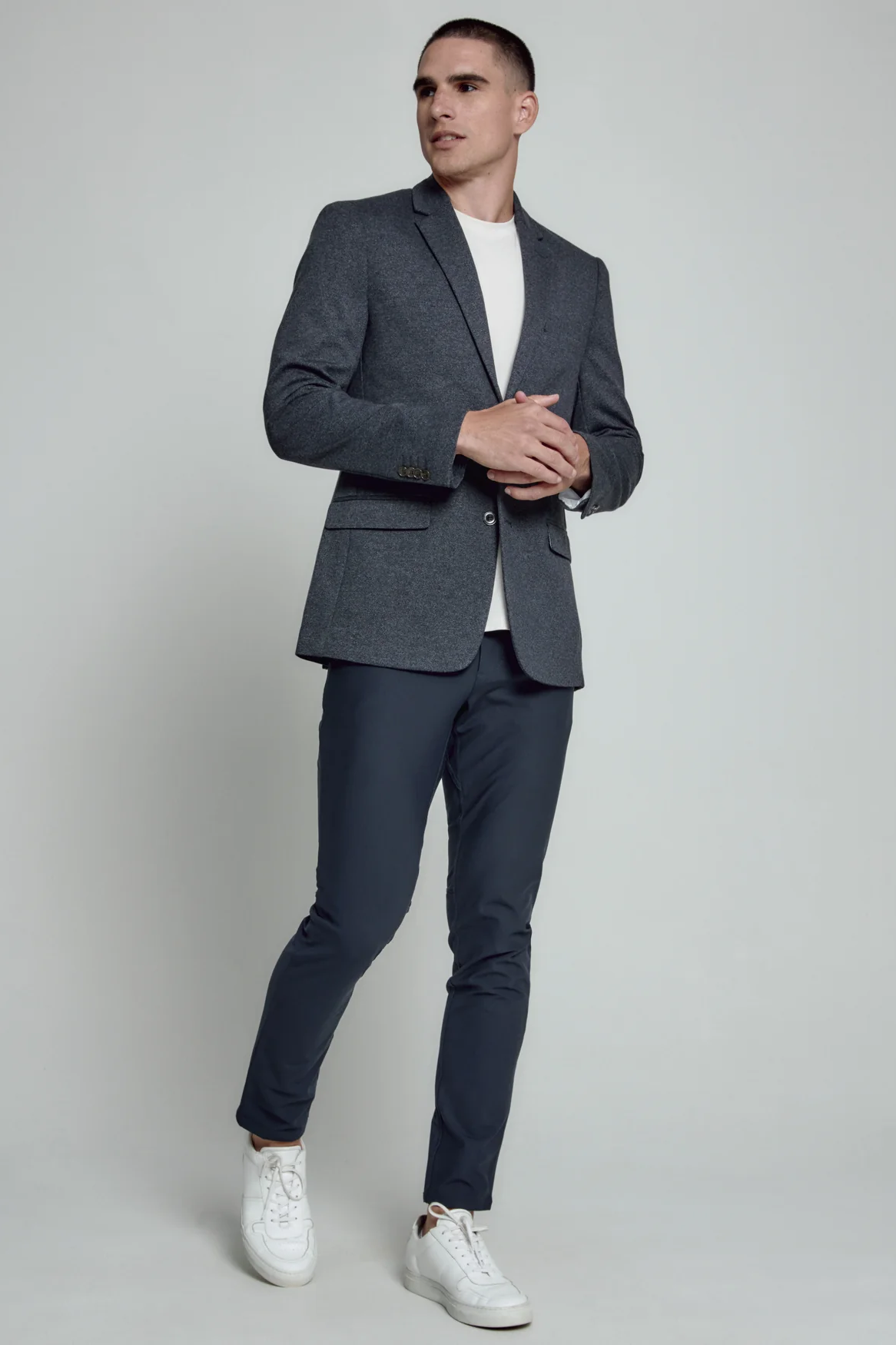 Man wearing a gray blazer, navy pants, and white sneakers on a plain background