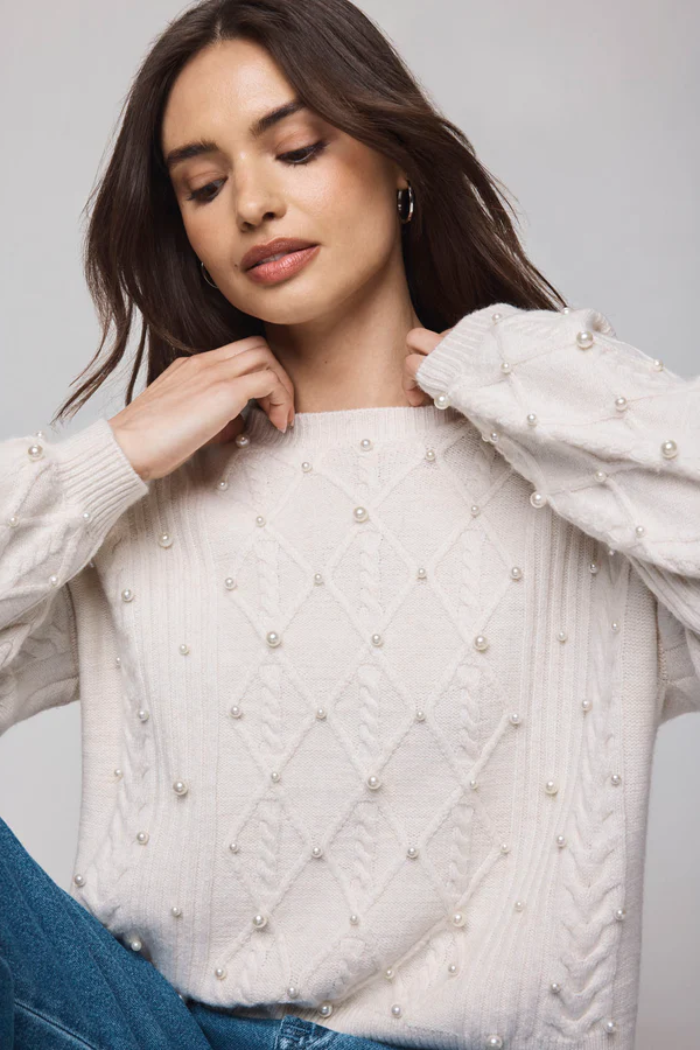 Woman wearing a white sweater with pearl embellishments against a plain background