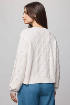 Woman wearing a cream-colored cable knit sweater and blue jeans on a gray background