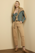 Woman wearing a patterned top and beige pants against a beige wall.