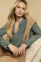 Woman wearing a green polka dot blouse and beige vest against a beige background