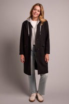 Woman wearing a black coat over a white hoodie, standing against a plain background