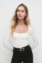 Woman wearing a white long-sleeve top and black pants with a gold belt against a white background