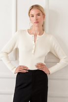 Woman wearing a white long-sleeve top and black pants against a white wall